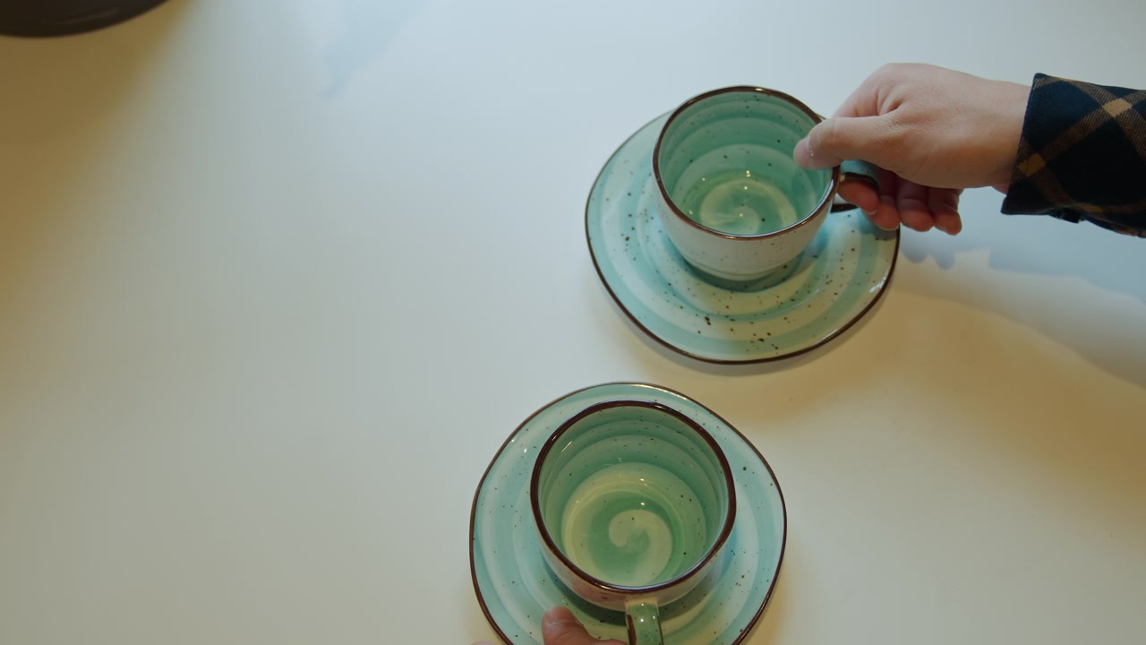 Close up of pair of hands carefully picking up matching ceramic cups and saucers with turquoise glaze from white table. sequence shows deliberate movement as each cup is lifted revealing