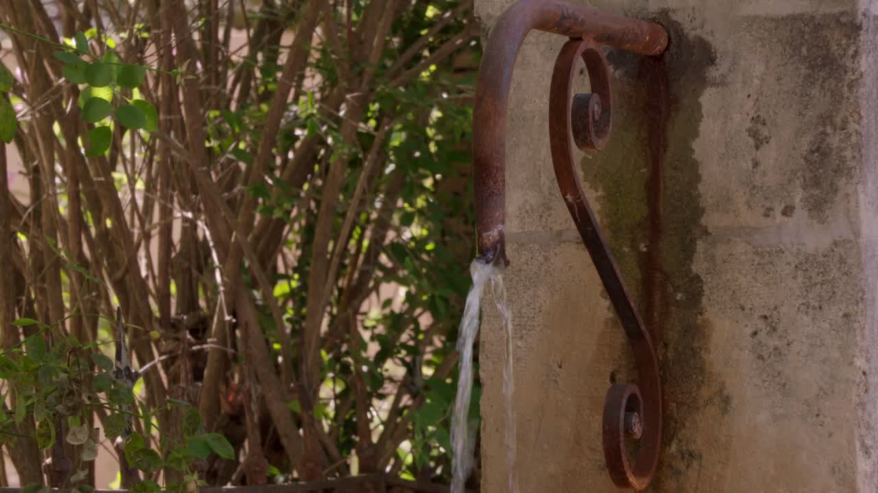 close-up shot of a rustic, weathered stone wall in Provence, France, with a rusted metal pipe faucet dispensing a stream of water, surrounded by greenery.