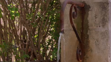 close-up shot of a rustic, weathered stone wall in Provence, France, with a rusted metal pipe faucet dispensing a stream of water, surrounded by greenery.