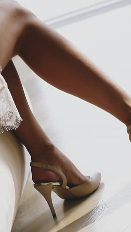 cu shot, slow motion: slim legs of graceful young woman wearing stylish white lacy rest gown and high heeled shoes sitting on bed in bedroom close view