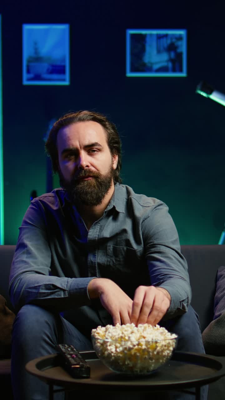 Vertical video Man enjoying bowl of popcorn and smiling while watching cable programs on TV