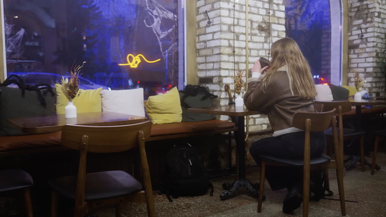 Beautiful girl in brown shearling jacket sitting at cozy cafe sipping coffee near window with people walking outside in soft natural lighting creating warm intimate atmosphere