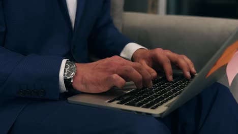 Close-up-of-businessman-hands-typing-on-laptop-keyboard-while-sitting-on-sofa-holding-grey-laptop-on-knees.-A-man-wearing-blue-formal-suit-and-expensive,-fancy-watches