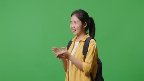 side view of asian woman student with a backpack and some books smiling and clapping her hands while standing in the green screen background studio