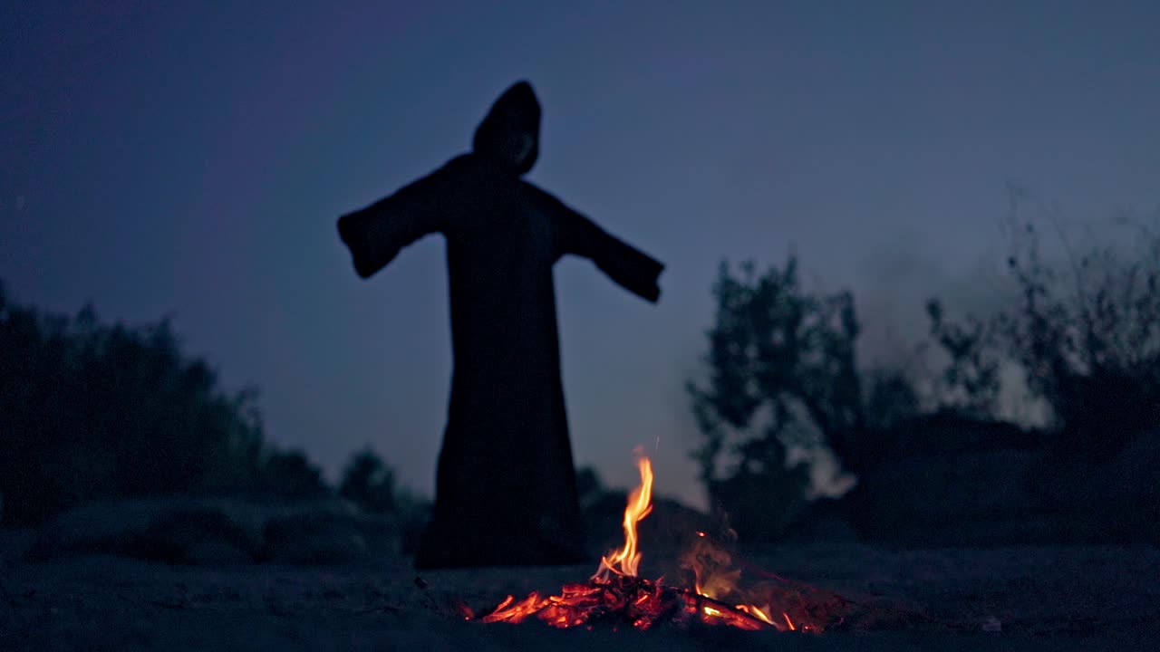 Black wizard or witch going around the bonfire outdoors. Silhouette of death performing some ritual near the fire in the evening.