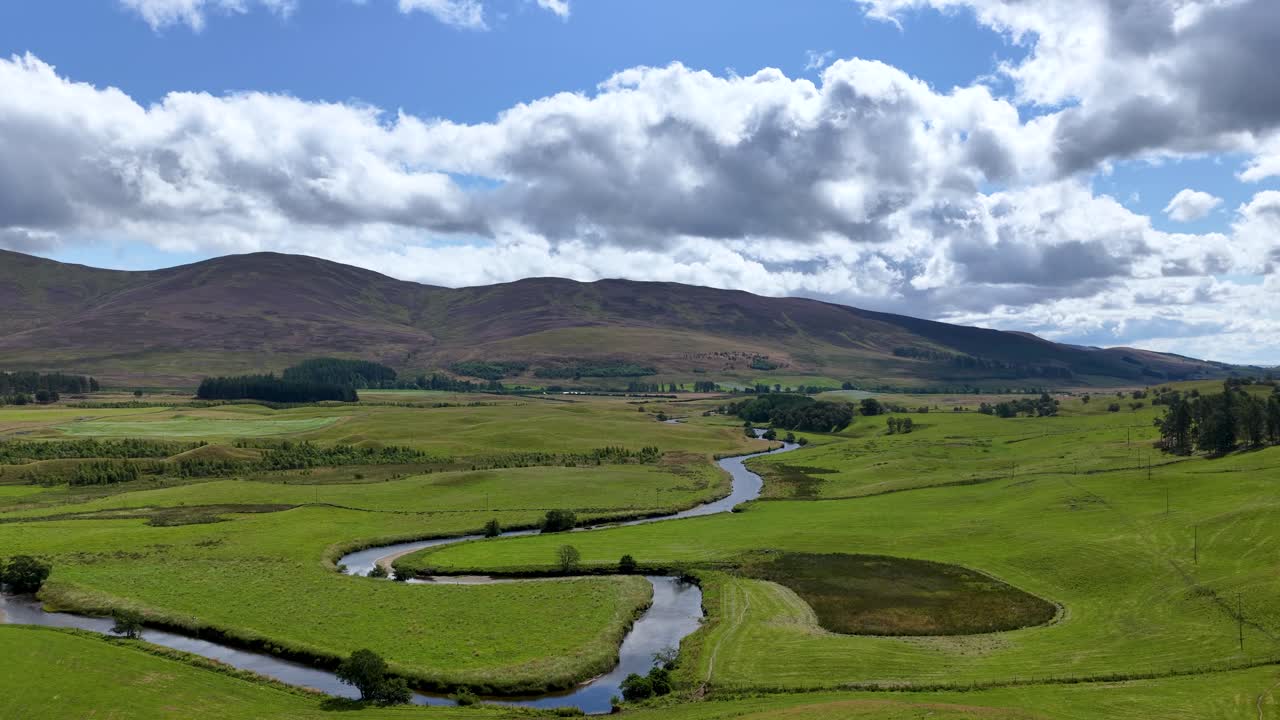 Drone glides above lush green meadows, winding river, and hills under partly cloudy daylight