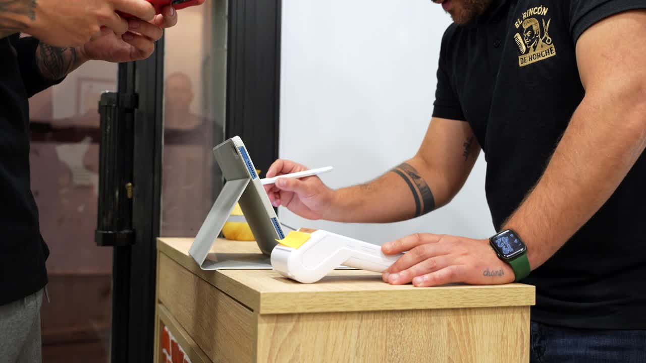 Handheld shot of customer placing phone over dataphone for contactless payment while barber confirms transaction on tablet with stylus