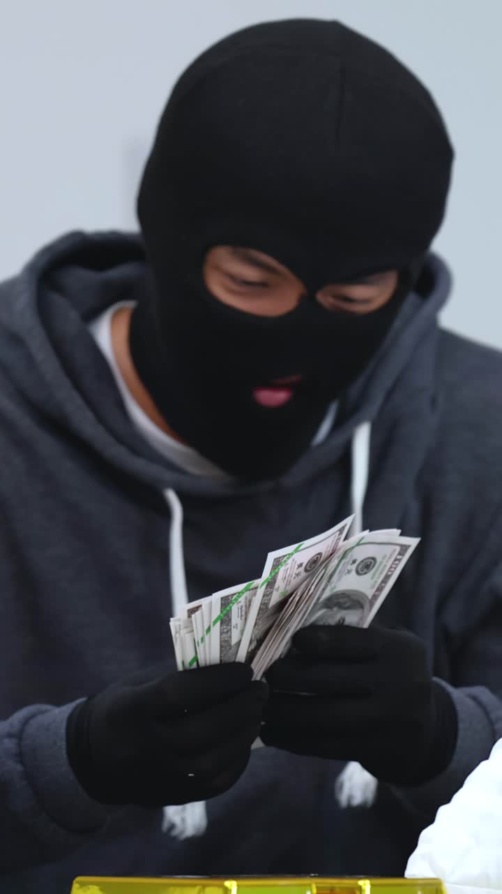 Un criminale con un passamontagna conta mazzette di dollari sopra lingotti d'oro, all'interno.