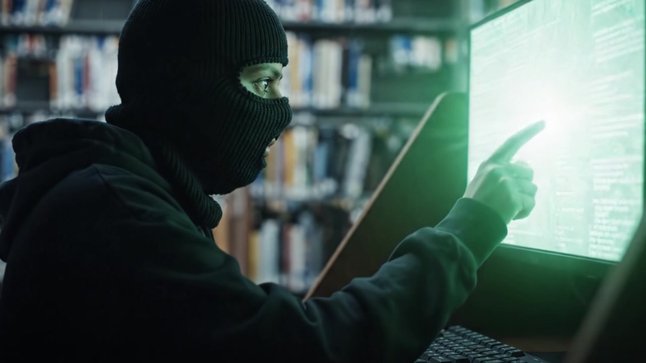 Un individuo mascherato si impegna in un'intrusione informatica in una biblioteca, ottenendo accesso non autorizzato a informazioni riservate su un terminale informatico.