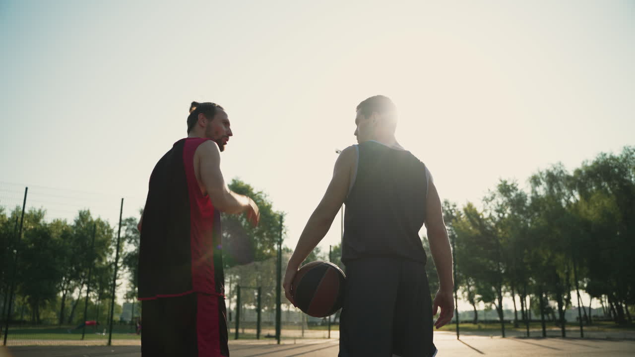 Back View Of Two Male Basketball Players Holding Balls And Walking Away From Camera, While Talking Each Other In An Outdoor Basketball Court