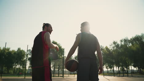 Vista-Trasera-De-Dos-Jugadores-De-Baloncesto-Masculinos-Sosteniendo-Pelotas-Y-Alejándose-De-La-Cámara,-Mientras-Hablan-Entre-Ellos-En-Una-Cancha-De-Baloncesto-Al-Aire-Libre