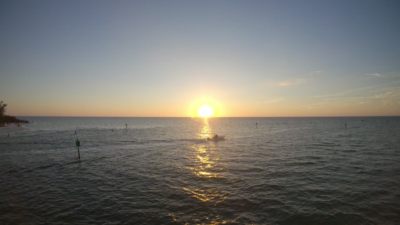 A small boat sailing out from a beach in Naples, Florida with the sun setting over the sea.