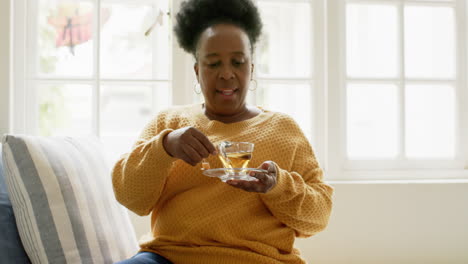 Feliz-Mujer-Mayor-Afroamericana-Sentada-Con-Té-Y-Sonriendo-En-Una-Habitación-Soleada,-Cámara-Lenta