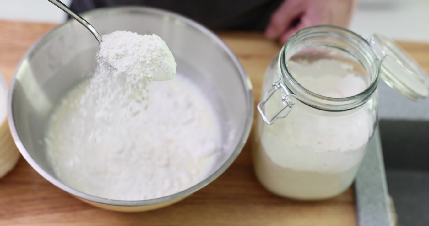Cuoco aggiunge farina in una grande ciotola di acciaio inossidabile in cucina.