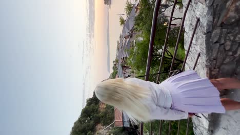 A-young-woman-dressed-in-a-white-shirt-and-purple-skirt-walks-down-the-staircase-to-the-beach-club-with-a-sunset-over-the-beautiful-Adriatic-ocean-in-Dubrovnik