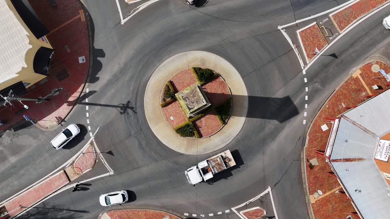 Overhead drone footage captures cars and trucks moving through a sunlit roundabout in a small Australian town, with steady camera and clear shadows