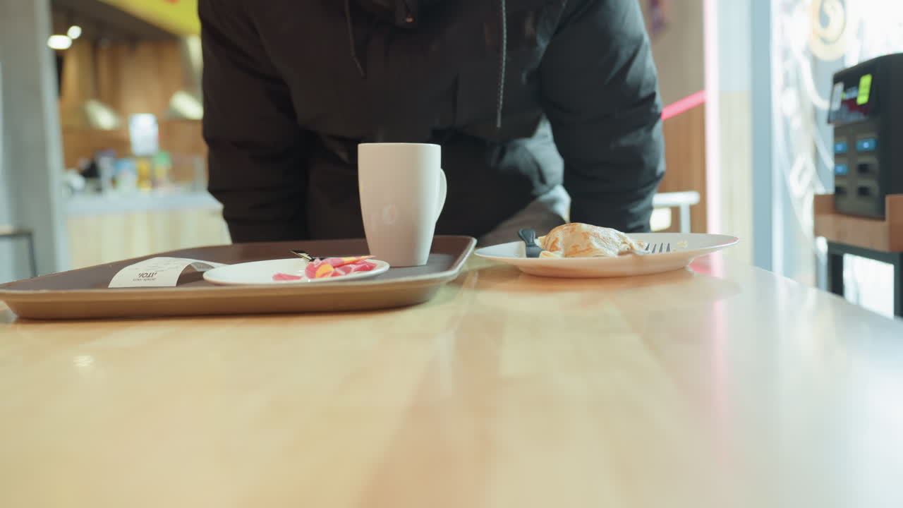 Tom bakke og krus på bordet, mand i mørk jakke nærmer sig og sætter bakke med kaffe og wienerbrød på træbord, varmt dagslys, blød bokeh-baggrund af travl cafeteria, spontant nærbillede der formidler.
