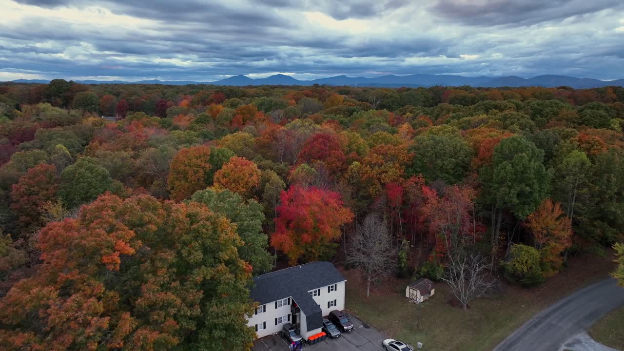 Aerial flyover American suburban neighborhood surrounded by vibrant autumn foliage, colorful trees and distant mountain ranges under dramatic clouds. Virginia, United States in October