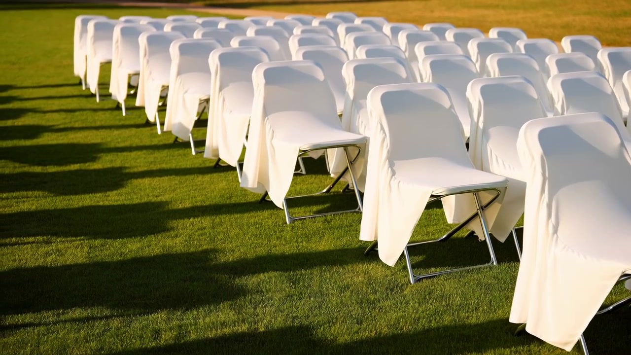 Low oblique view of neatly aligned metal framed folding chairs draped in white fabric across manicured grass, creating repetition, depth, and warm late afternoon shadow patterns, with chair, rows