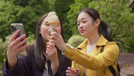 Two-Smiling-Young-Female-Friends-Meeting-And-Posing-For-Selfie-Eating-Ice-Cream-Outdoors-In-Park-Together-1