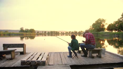 camera zooming from the back of a teen boy sitting with his grandfather on the lake pier, talking and fishing together