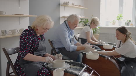 medium shot of middle aged ceramic artist teaching group elderly caucasian woman and senior man how to wedge clay sitting at desk in art studio. people enjoying talking at work