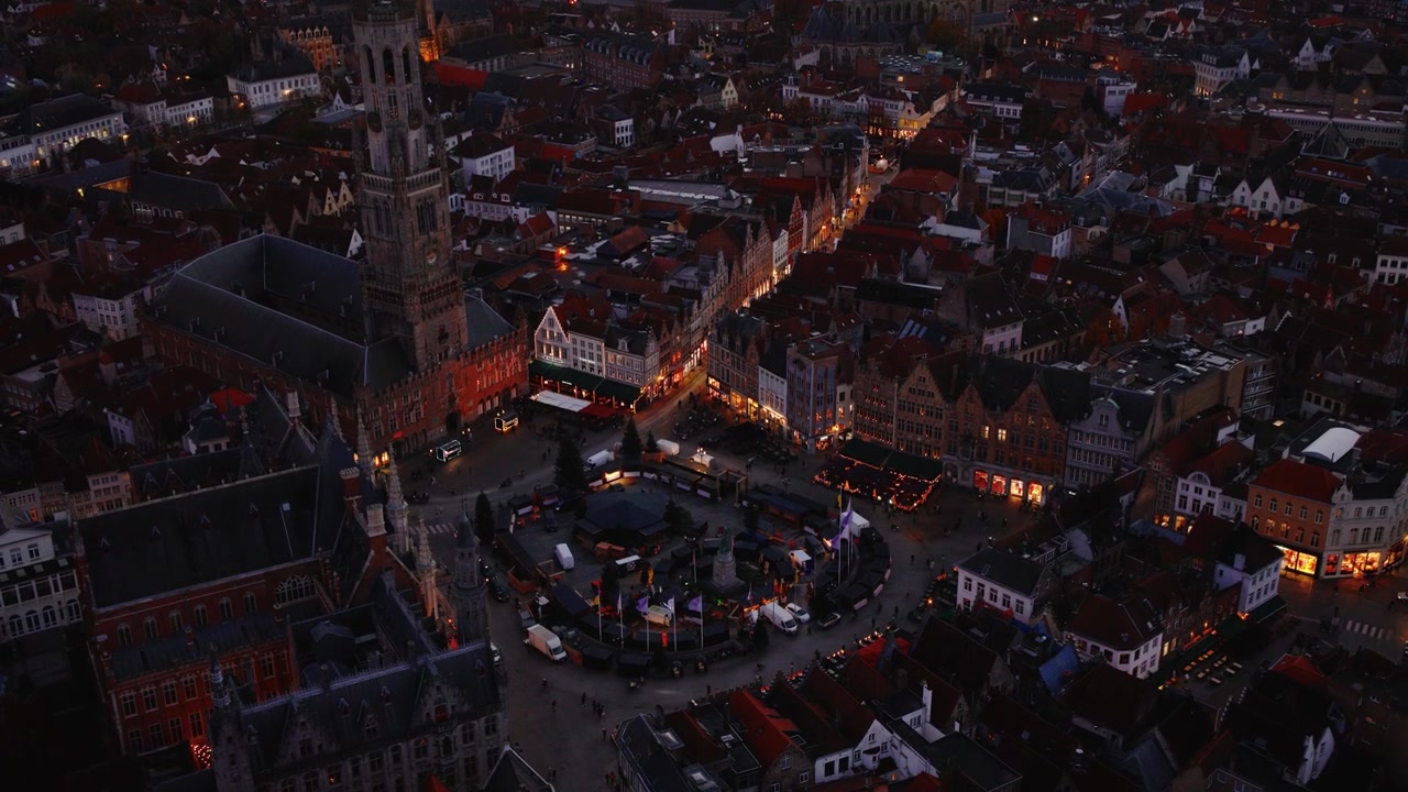 Filmato drone che inizia con un'ampia panoramica di Bruges e si sposta verso il Grote Markt, mostrando il centro storico, l'illuminazione serale e gli edifici illuminati.