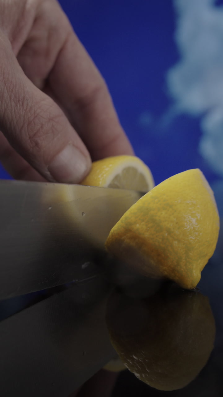 Vertical 9:16 slow motion video of a knife slicing a fresh yellow lemon on a wet surface while clouds drift through a blue sky behind