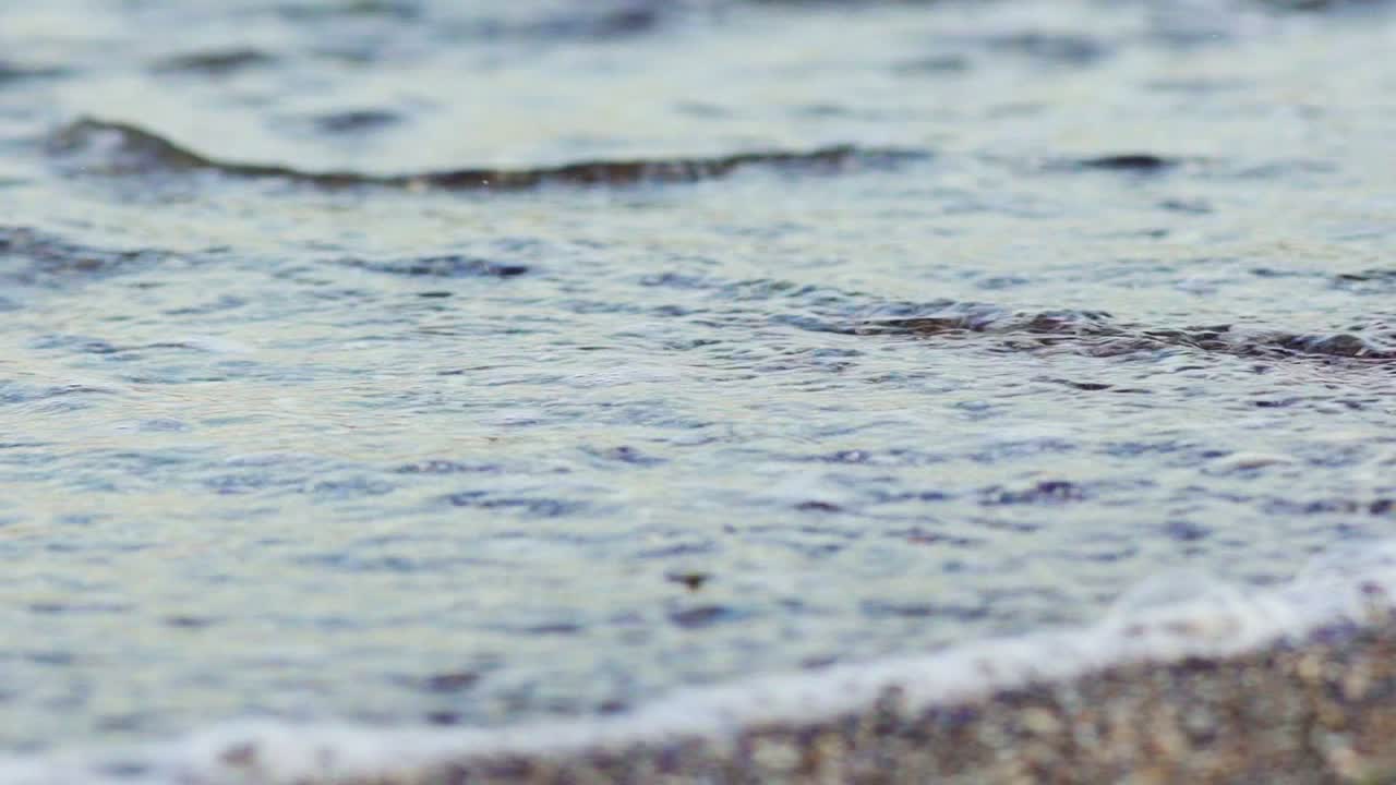 Flowing sea water on the pebble beach in the evening. The edge of the ocean with calm waves on the pebble background. Close-up.