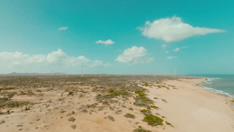Luftaufnahme-Des-Wüstenstrandes-Und-Des-Ozeans-Mit-Windkraftanlagen-Im-Hintergrund,-Kolumbien,-La-Guajira