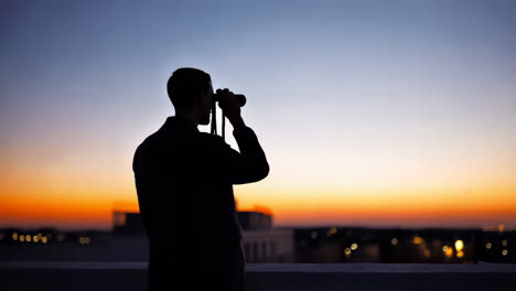 silhouette of a man looking through binoculars from a rooftop at sunset