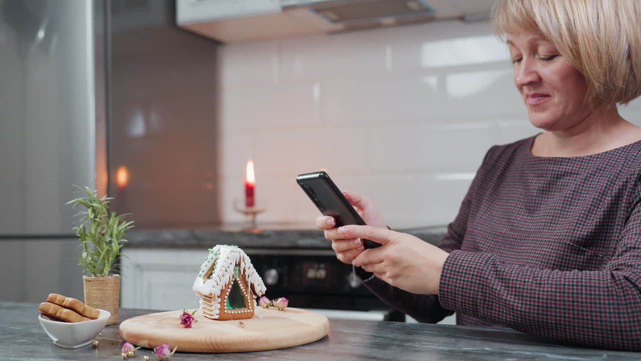 una mujer sonriente toma una foto de su casa de pan de jengibre decorada usando un teléfono inteligente, el entorno cálido de la cocina presenta galletas, plantas en maceta y equipos de cocina
