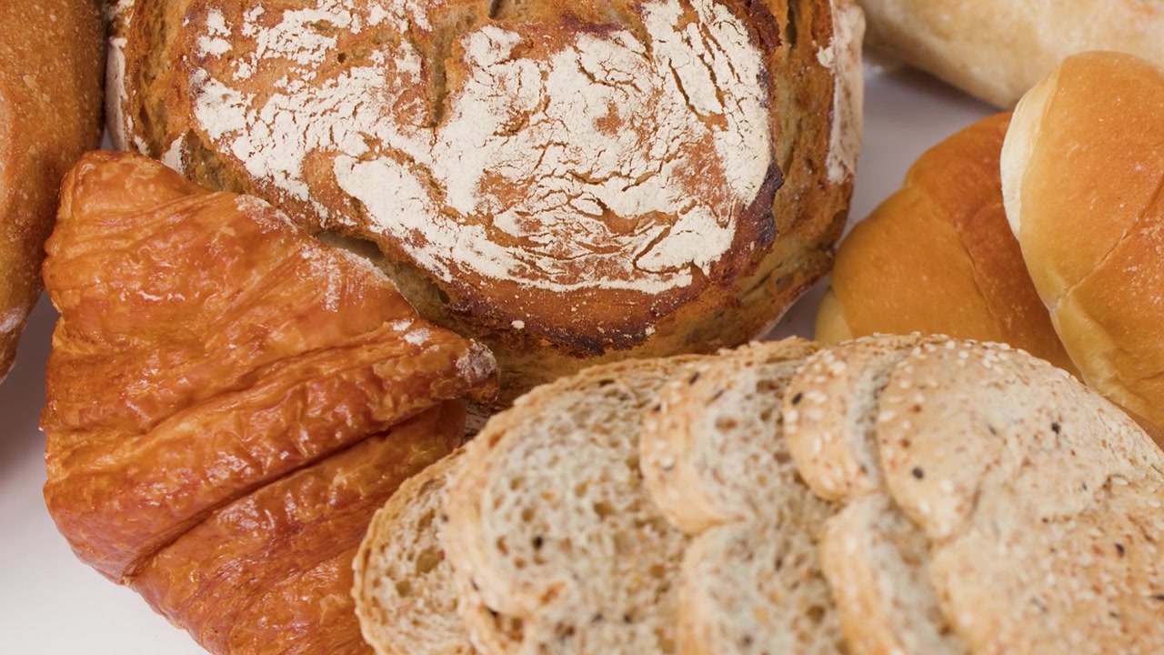 Slow zoom out reveals sourdough, croissants, and sliced whole grain bread on white background.