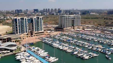 sailing yachts navigate into herzliya's marina as another sailboat departs from its berth in israel as several tourists stroll the boulevard