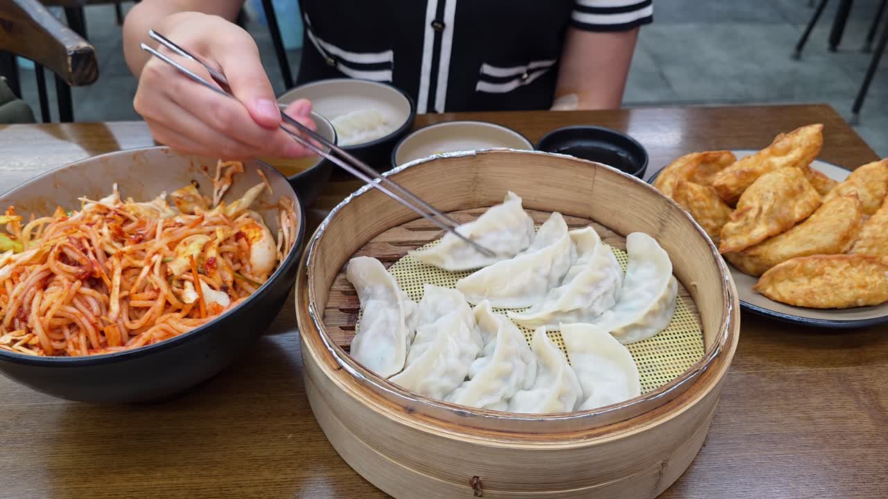 A woman uses chopsticks to pick up a delicious steamed mandu from a bamboo steamer, part of a traditional Korean meal that also includes fried dumplings and spicy noodles at an authentic restaurant