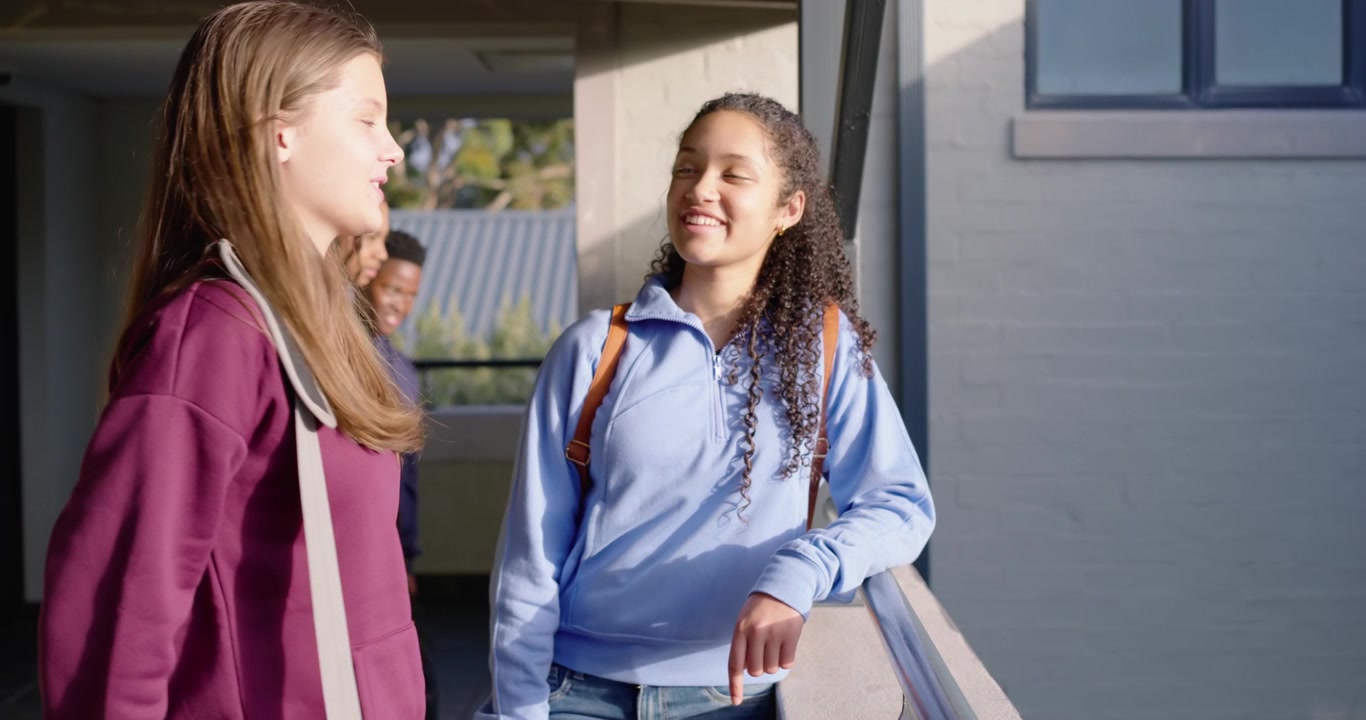 Teen females meeting after class on school balcony, in blue hoodie, leaning on railing, chatting