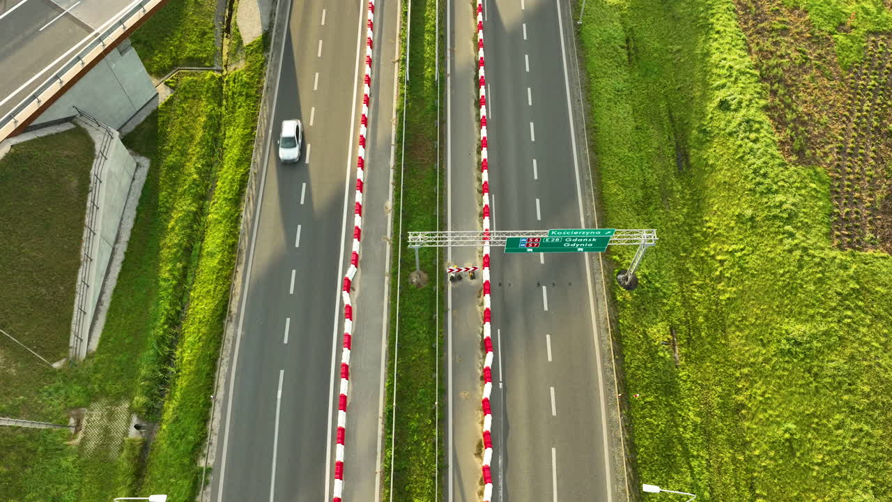 Close top-down drone view of the S6 expressway lanes, showing a white car passing beneath an overhead road sign gantry, framed by red and white barriers