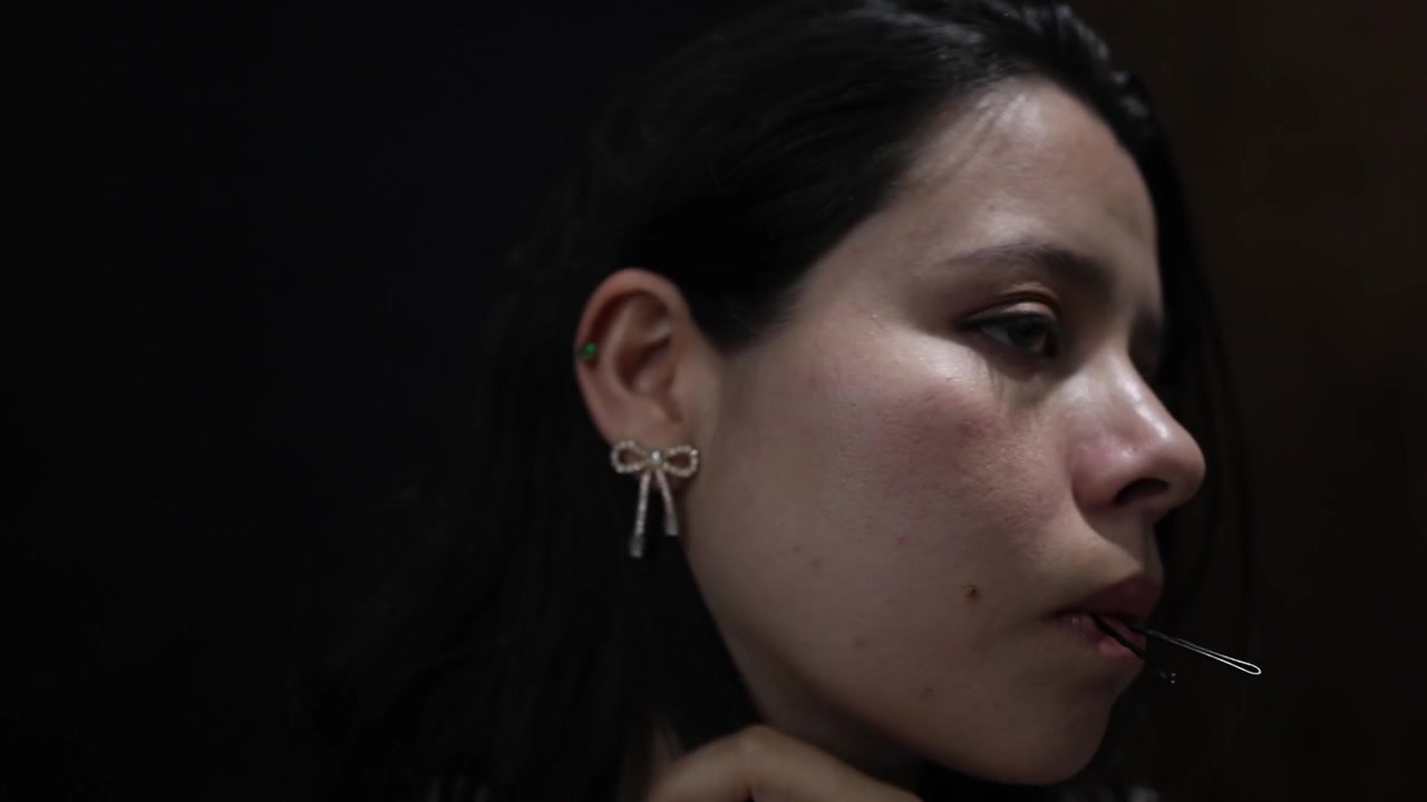 Primo piano di una giovane donna che si lega i capelli, mostrando intricati movimenti delle mani e tratti delicati. La scena cattura un momento di tranquilla concentrazione con un'illuminazione morbida e a basso contrasto.