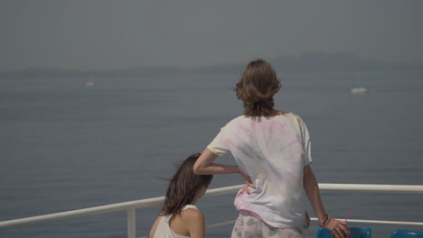 two young women tourist stand on the deck of a cruise ship and look out over the ocean while the boat is sailing. wind blows in their hair. adventure, freedom and lifestyle concept, 4k slow motion.