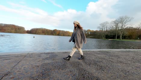 Glückliche-Frau,-Die-Während-Der-Herbstsaison-An-Einem-Bewölkten-Tag-In-Einem-Park-In-Der-Nähe-Eines-Sees-Spazieren-Geht