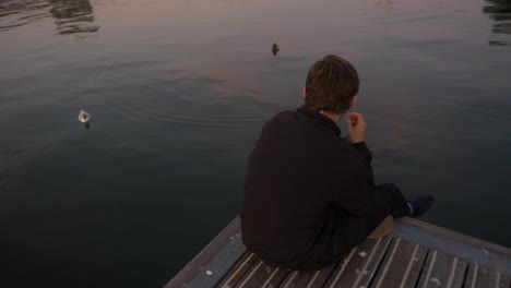 overhead view of a young man sitting on the edge of a lake pier eating potatoe chips and looking at the ducks below