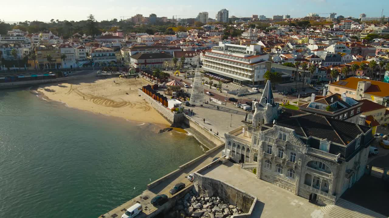 Vídeo aéreo grabado con dron del Palacio de Seixas en la playa de arena de Estroil, Cascais, Portugal, y la costa turística durante un día soleado, con apartamentos y casas de lujo visibles en las calles por donde pasean los turistas