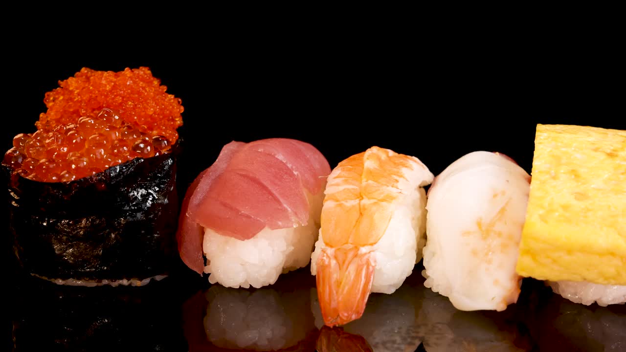 A variety of nigiri and sashimi, including eel, tamago, squid, shrimp, salmon roe, scallop, salmon, and tuna, rotate smoothly on a glossy black surface under bright studio lighting