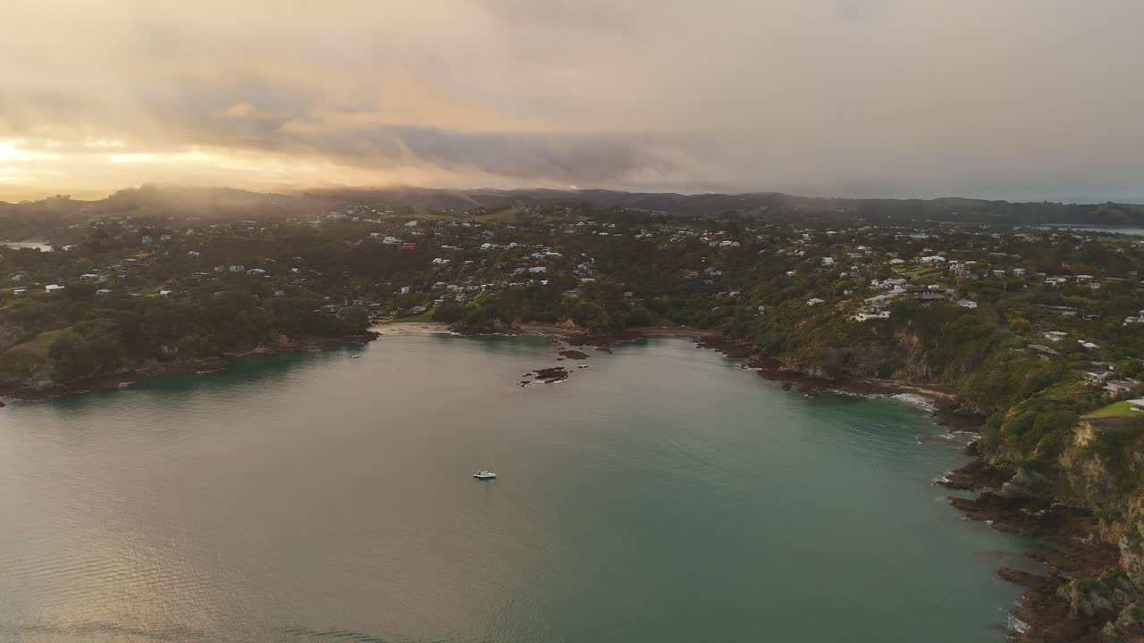 Aerial wide shot of sandy beach and bay of Waiheke Island with green mountains and flying clouds. Sunset time in New Zealand.