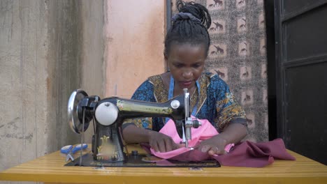 foto frontal de una mujer africana cosiendo telas en una máquina de sastrería en áfrica afuera de su pequeña casa en cámara lenta.