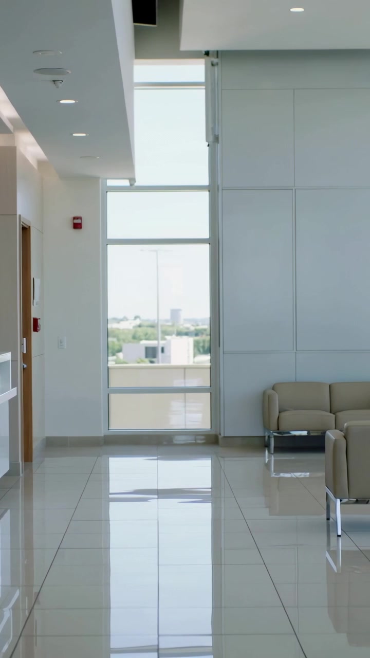 Vídeo vertical: Câmera panorâmica pela área do lobby mostrando um monitor, telefone, poltronas bege e uma janela.