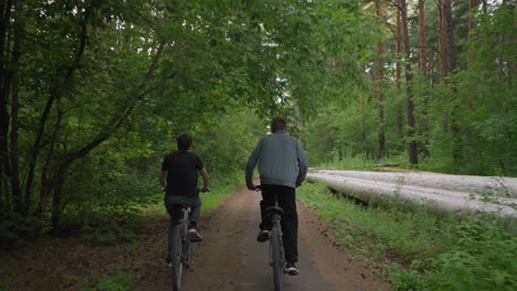 two friends riding bicycles along a paved untarred path surrounded by lush green trees, one of them stands while cycling, a log of wood lies along the side