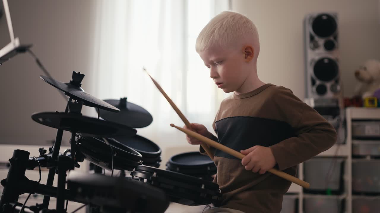 Shot from the side of a small albino boy with white hair and blue eyes playing and tuning his electric musical drum kit using special wooden sticks during his rest and day off in his room
