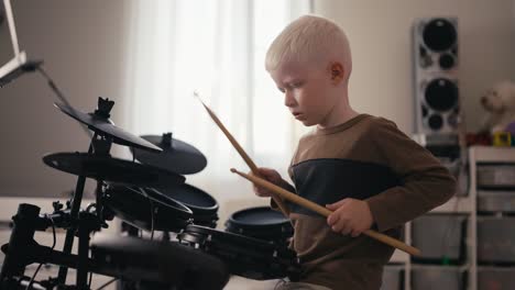 Shot-from-the-side-of-a-small-albino-boy-with-white-hair-and-blue-eyes-playing-and-tuning-his-electric-musical-drum-kit-using-special-wooden-sticks-during-his-rest-and-day-off-in-his-room