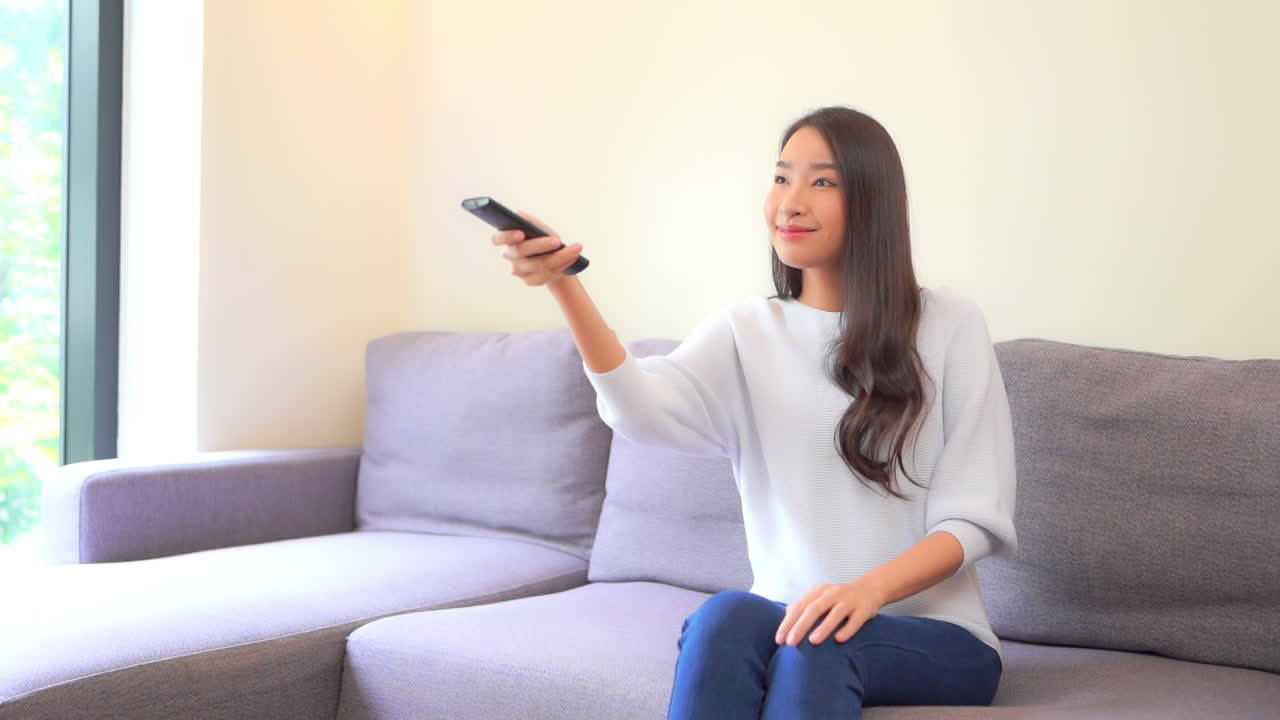 청바지와 스웨터를 입은 젊은 여성이 채널을 변경하기 위해 클릭하는 장치에서 리모콘을 가리킵니다.